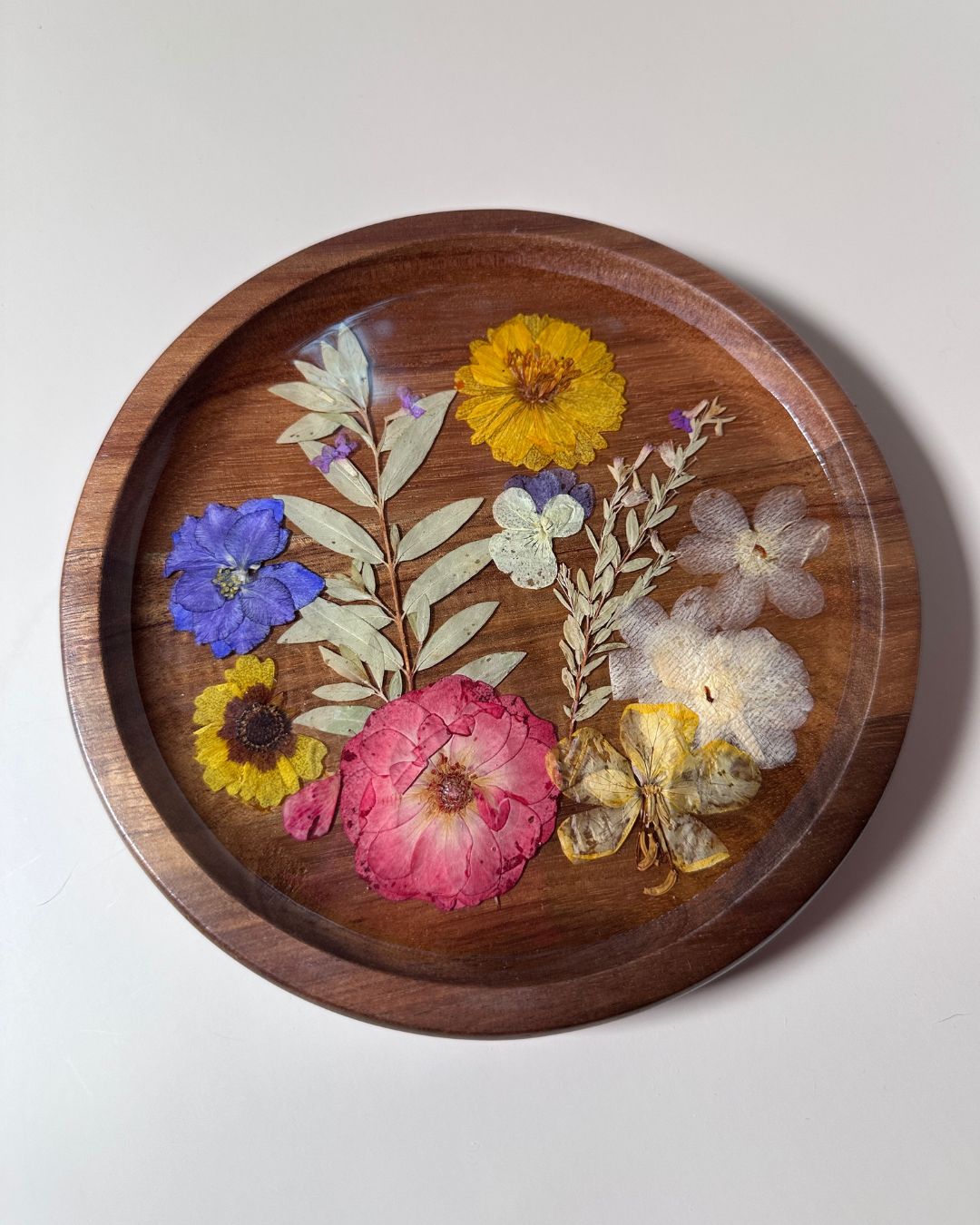 A round wooden tray with a clear top, filled with pressed dried and fresh flowers in pink, yellow, blue, and white, set against a plain light gray background.