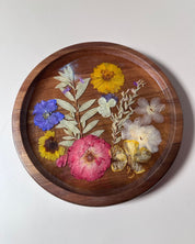 A round wooden tray with a clear top, filled with pressed dried and fresh flowers in pink, yellow, blue, and white, set against a plain light gray background.