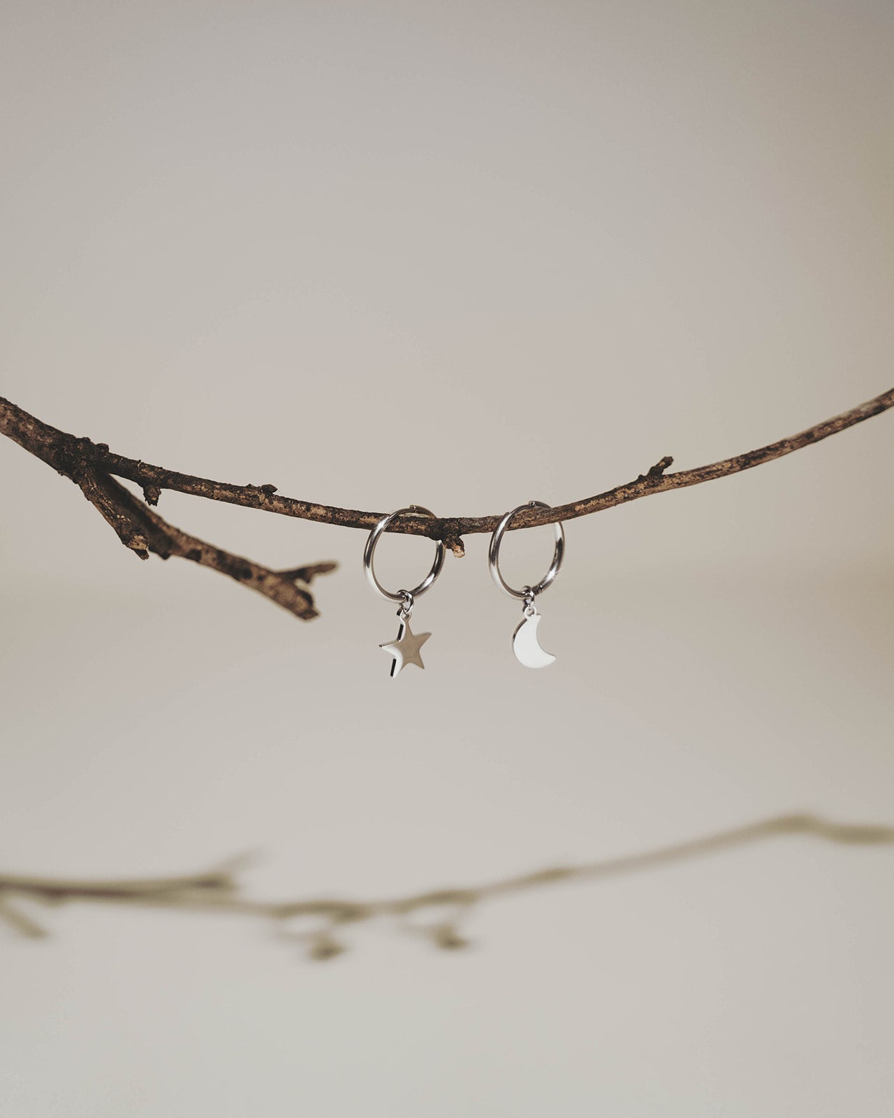 A pair of silver-toned hoop earrings, one with a small star charm and the other with a crescent moon charm, hanging from a thin brown twig against a soft beige background.