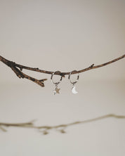 A pair of silver-toned hoop earrings, one with a small star charm and the other with a crescent moon charm, hanging from a thin brown twig against a soft beige background.
