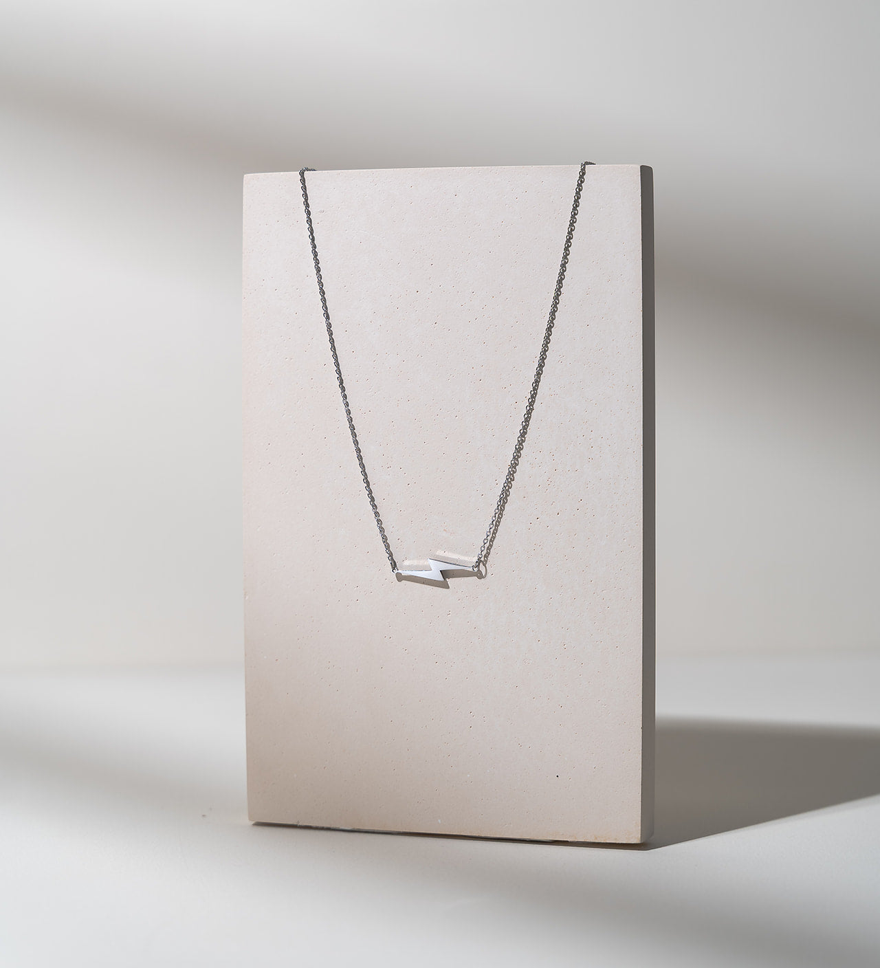 A delicate silver necklace with two small horizontal bar elements, displayed on a minimalist beige rectangular stand against a light gray background.