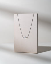 A delicate silver necklace with two small horizontal bar elements, displayed on a minimalist beige rectangular stand against a light gray background.