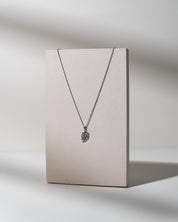 A silver-toned necklace with a small textured pendant displayed on a vertical beige card against a light gray background.