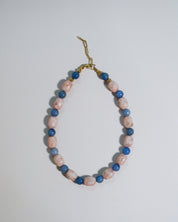 A beaded necklace with alternating light pink and medium blue polished beads, accented by small gold spacer beads and a gold clasp, shown on a plain light gray background.
