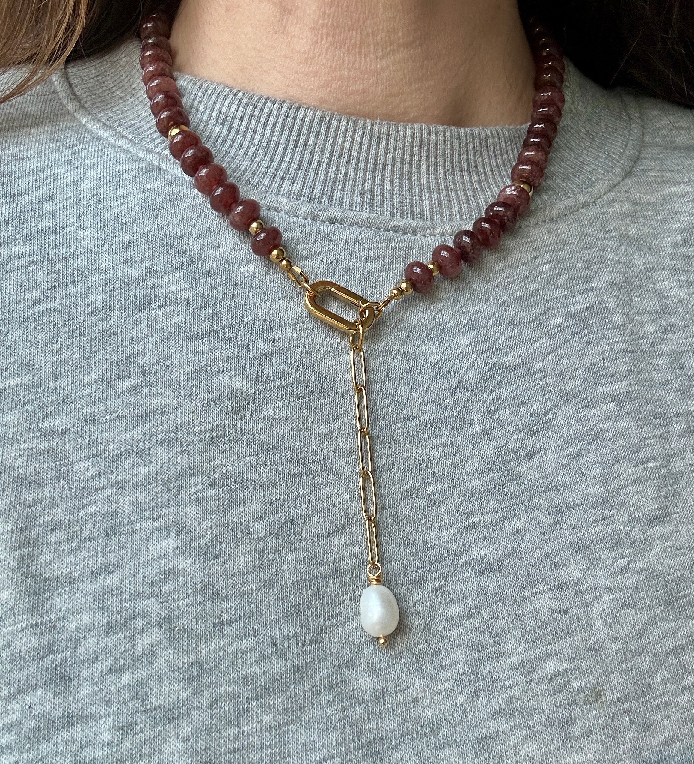 A close-up of a person wearing a beaded necklace with dark reddish-brown beads, a gold clasp, and a dangling white pearl-like bead on a gray knit top.