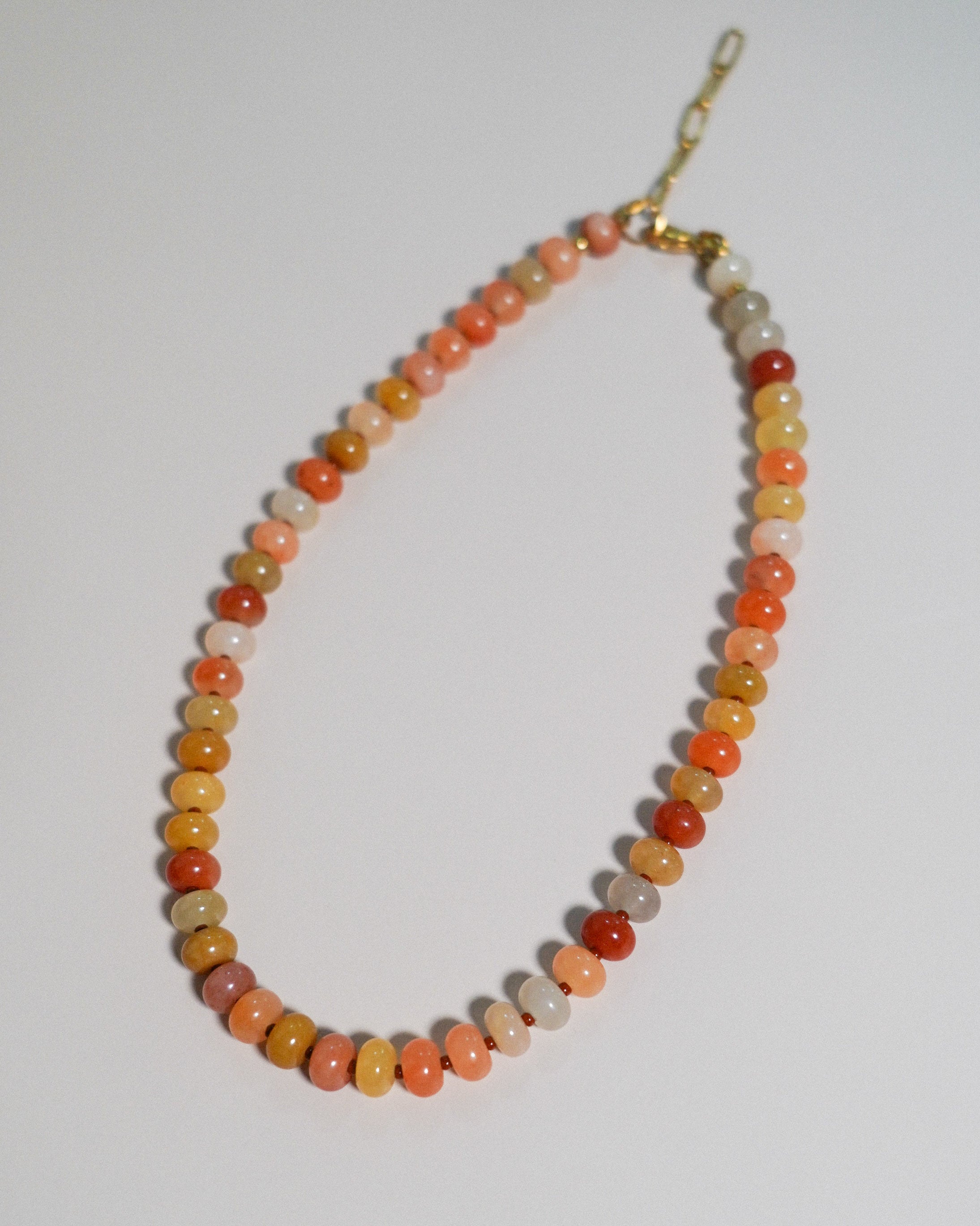 A beaded necklace made of small, polished oval beads in shades of orange, red, yellow, and cream, with a gold clasp, displayed on a plain light background.