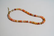 A beaded necklace made of small round stone beads in orange, peach, yellow, white, and brown, with a gold clasp, photographed on a plain light background.