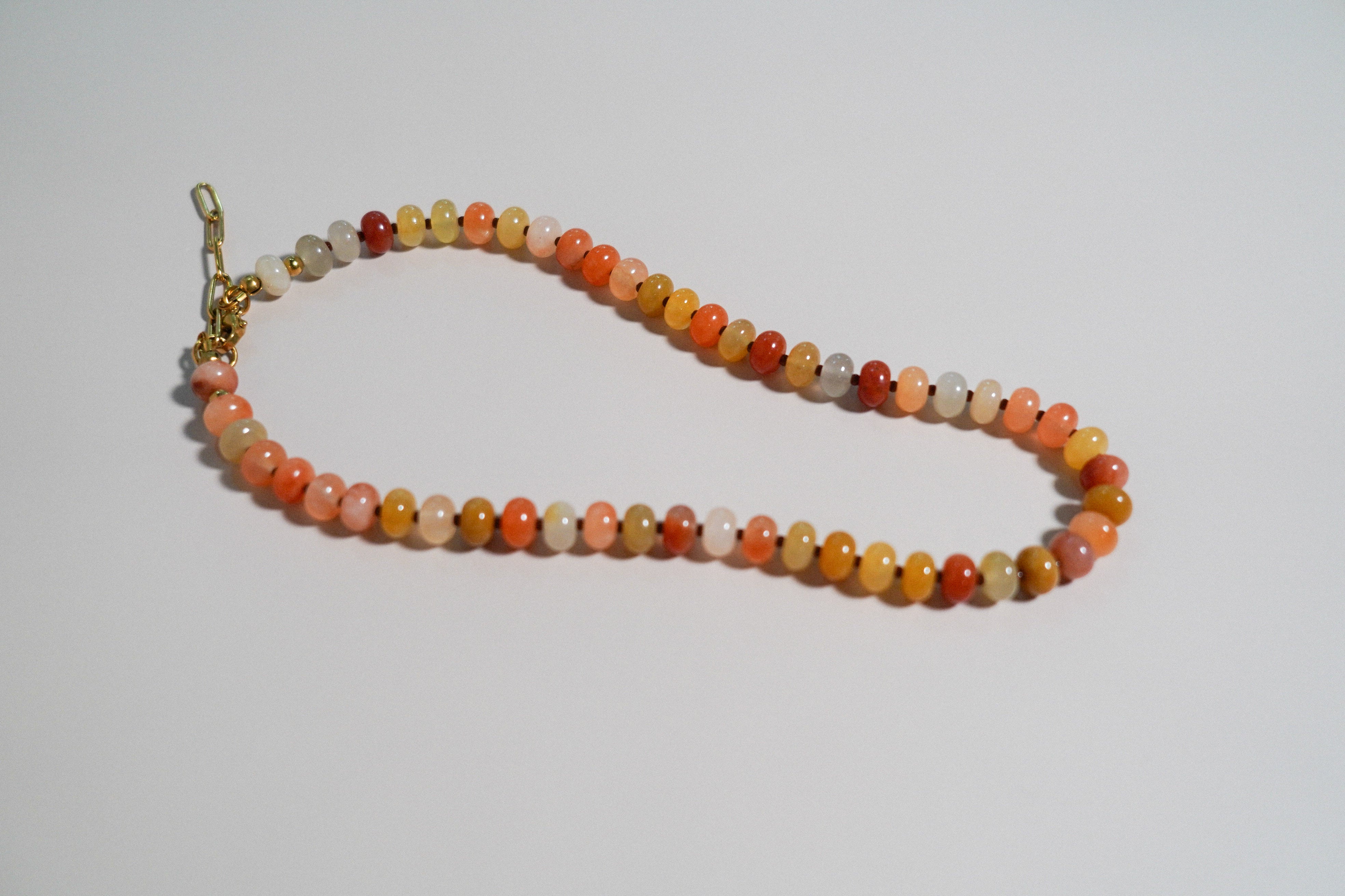 A beaded necklace made of small round stone beads in orange, peach, yellow, white, and brown, with a gold clasp, photographed on a plain light background.