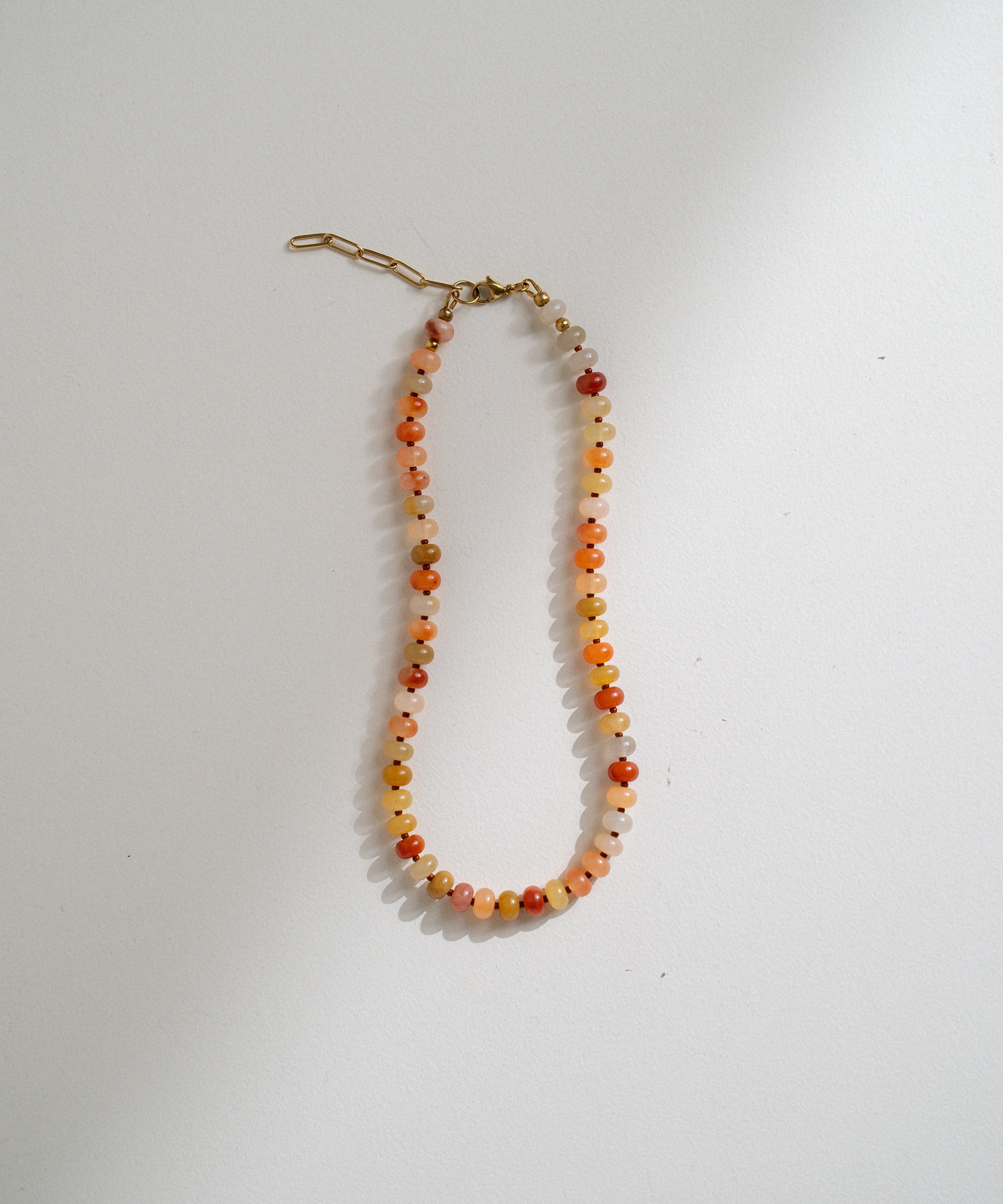 A beaded necklace with small round beads in orange, red, yellow, and white, featuring a gold-toned lobster clasp, displayed on a plain white surface.