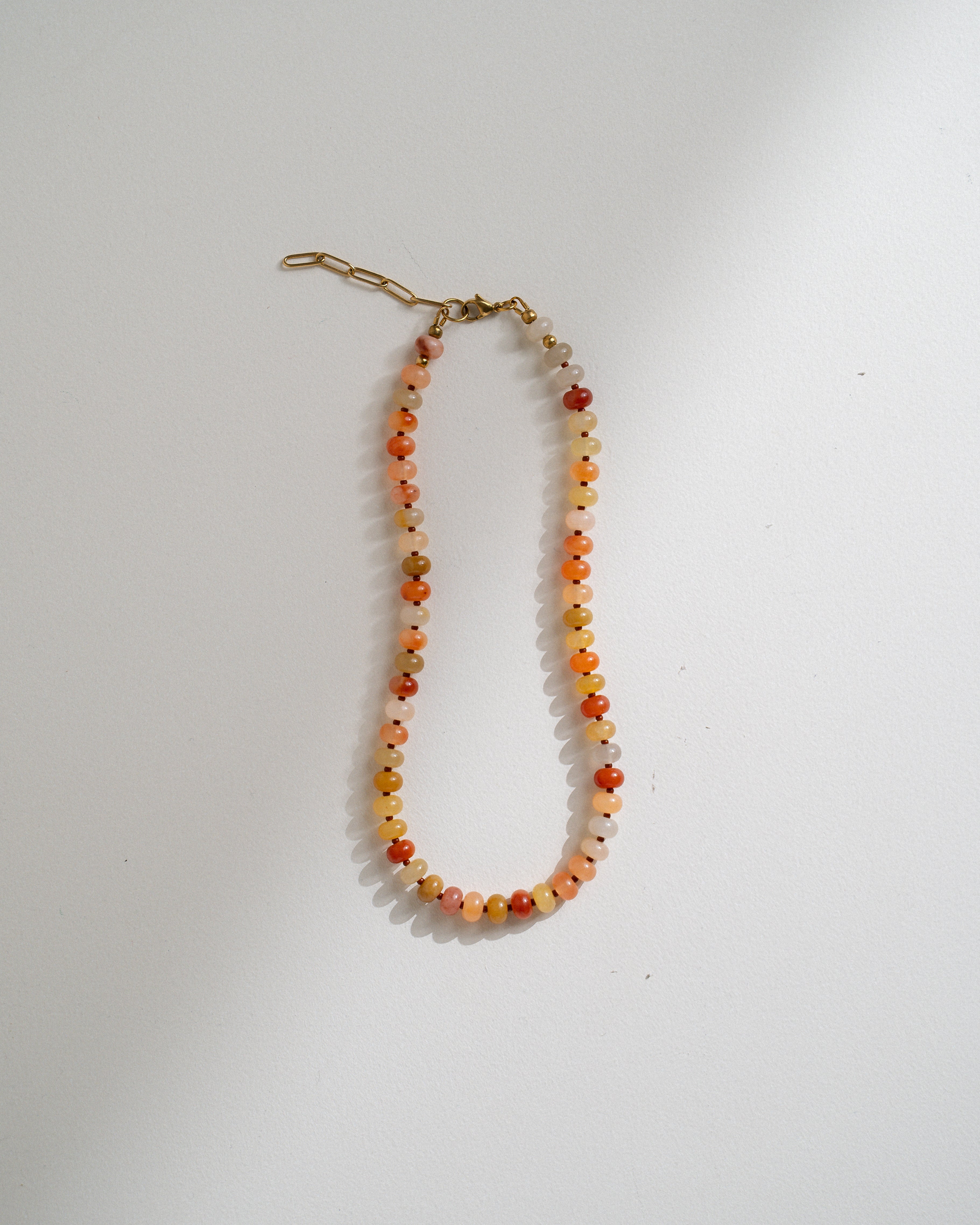A beaded necklace with small round beads in orange, red, yellow, and white, featuring a gold-toned lobster clasp, displayed on a plain white surface.