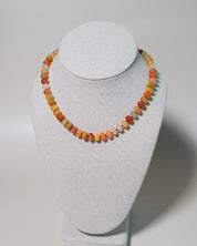 A beaded necklace made of small round gemstone beads in warm orange, yellow, peach, and cream tones, displayed on a light gray fabric jewelry bust against a neutral light gray background.