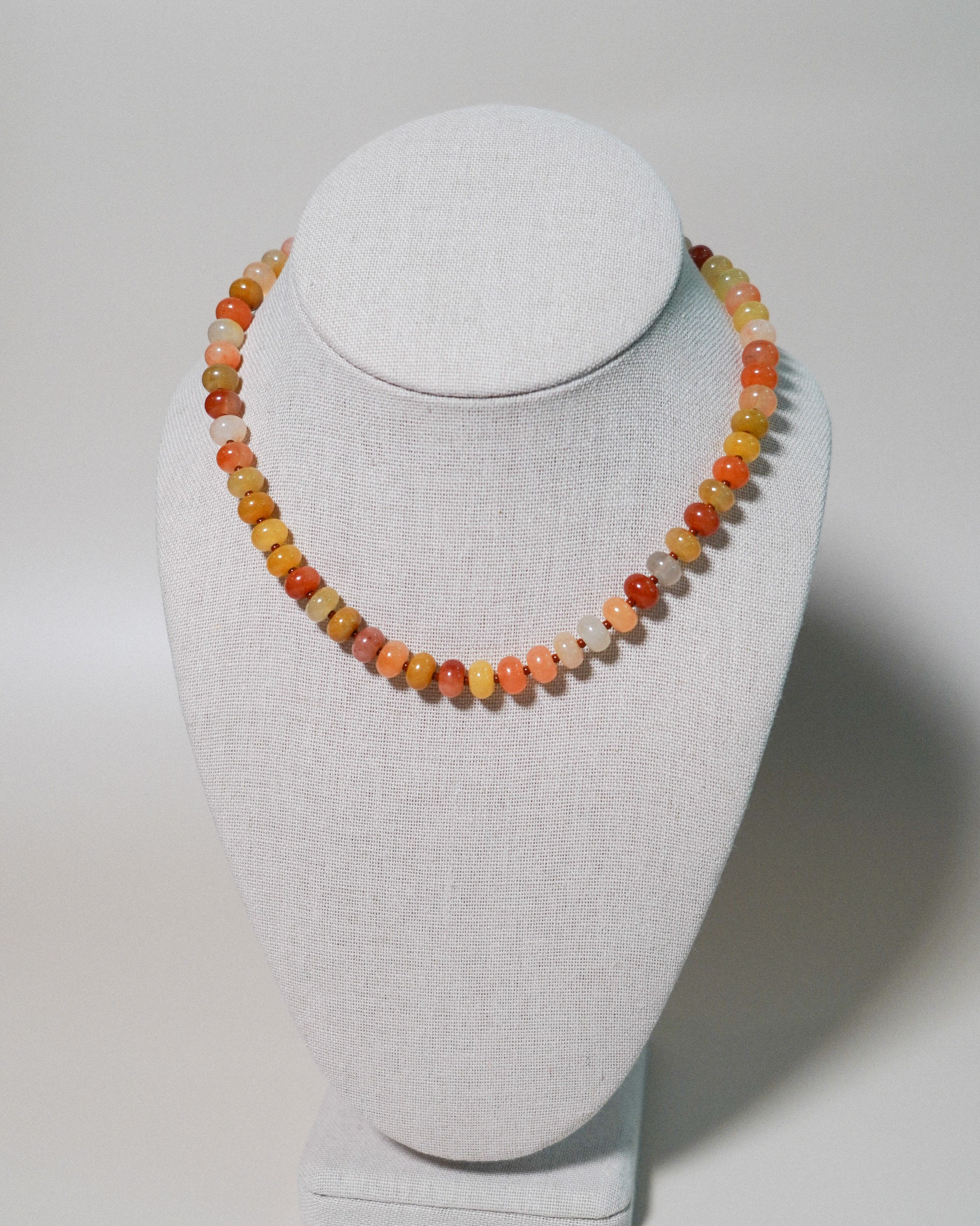 A beaded necklace made of small round gemstone beads in warm orange, yellow, peach, and cream tones, displayed on a light gray fabric jewelry bust against a neutral light gray background.