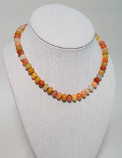 A beaded necklace made of small round polished stones in warm autumn colors, displayed on a white fabric-covered jewelry bust against a light neutral background.