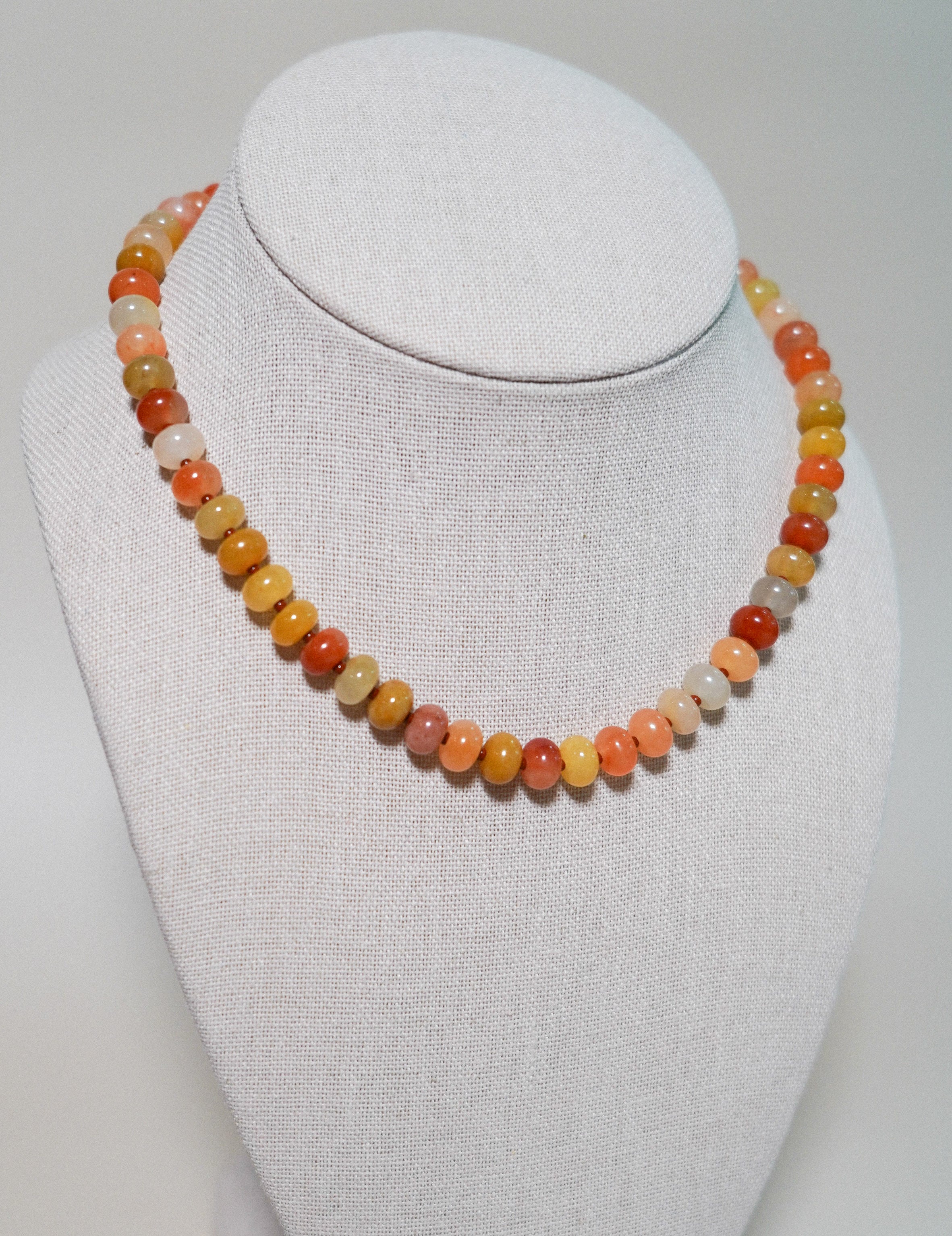 A beaded necklace made of small round polished stones in warm autumn colors, displayed on a white fabric-covered jewelry bust against a light neutral background.